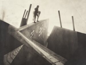 A still image from German expressionist classic The Cabinet of Dr. Caligari. The image depicts wildly distorted buildings where a male figure carries a woman across the roofs of the buildings.