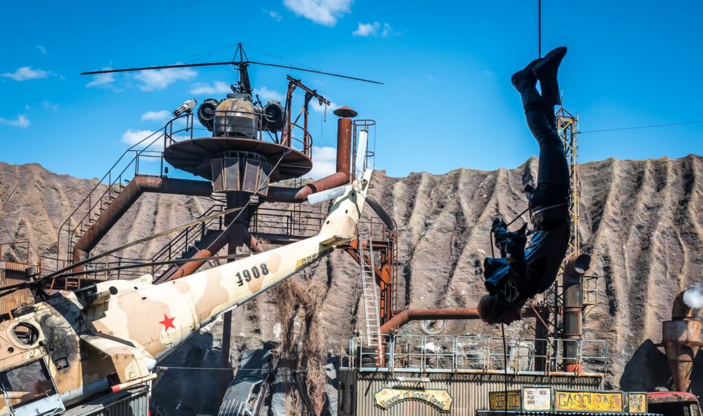 An image taken by Sergey Galyonkin at Filmpark Babelsberg. The image shows a stunt scene with a person descending mission-impossible style in the foreground; in the background there is a helicopter, motorbikes and junk.