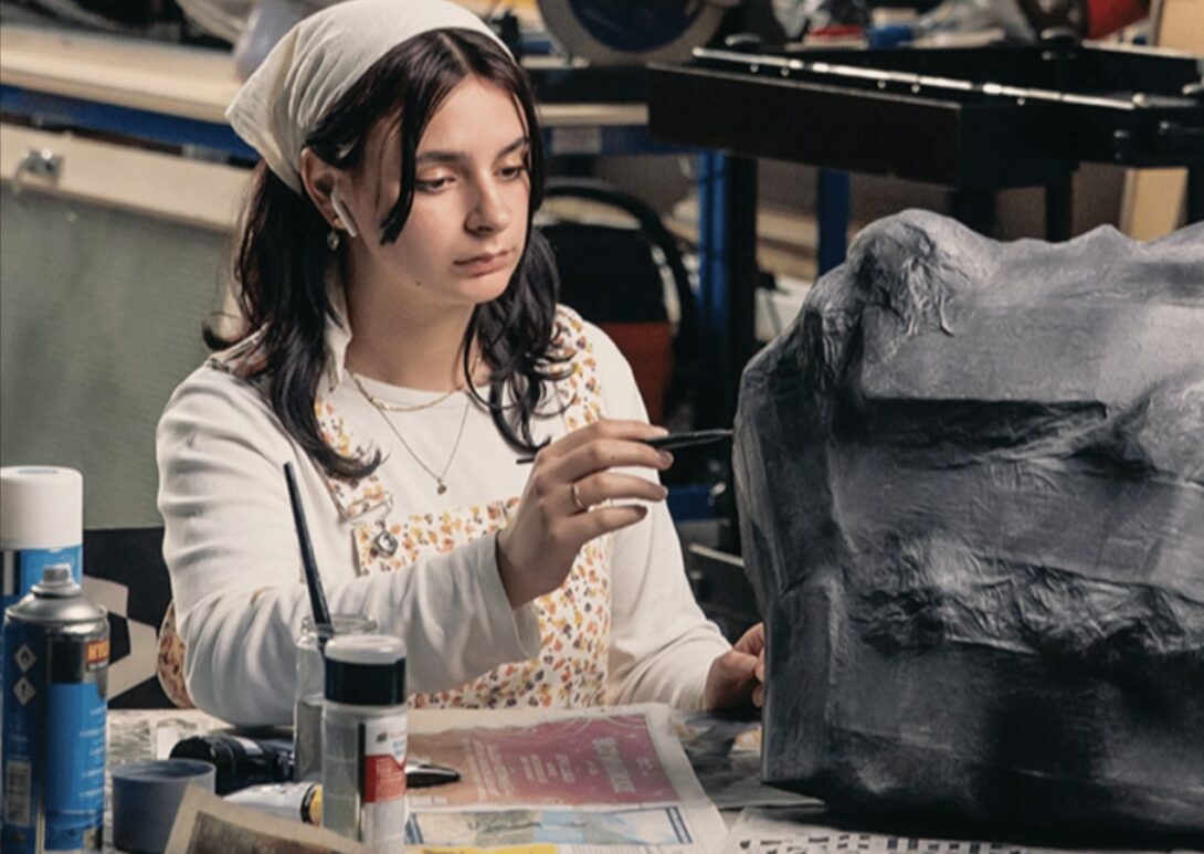 A student carefully painting a large prop in a workshop, surrounded by art tools and materials.