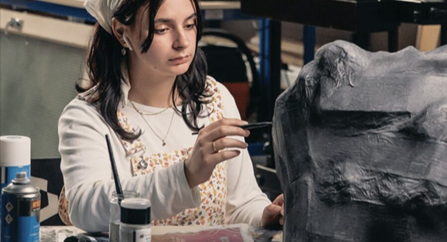 A student carefully painting a large prop in a workshop, surrounded by art tools and materials.